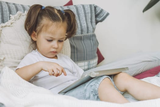 Little dark-haired girl 2-4 with two ponytails sits on bed with pillows playing tablet computer
