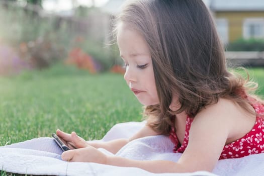 Portrait of little girl 3-4 in red clothes lying on blanket on green grass and looking into mobile phone. Children using gadgets