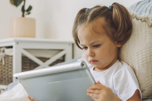 Little dark-haired girl 2-4 with two ponytails sits on bed with pillows seriously looking at tablet computer