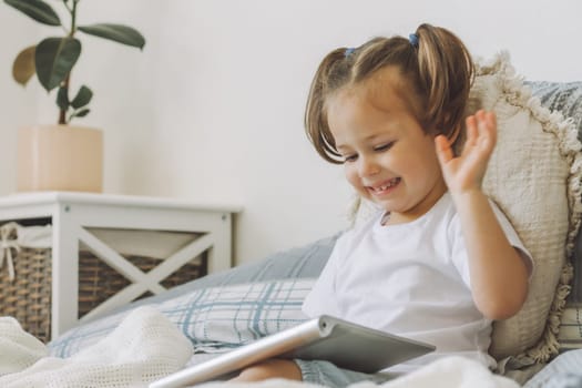 Little dark-haired girl 2-4 with two ponytails sits on bed with pillows and looks at tablet computer, smiles and waves her hand