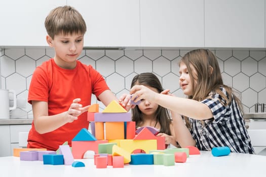 Positive, focused children of boy and two girls playing with colorful blocks and building castle from geometric figures on white table. Creative and art games. Motor activity and brainstorming game