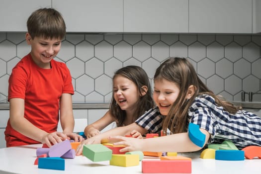 Laughing, funny and playful children of girls fooling around and boy playing with colorful bricks and building elements, throwing, scattering blocks on white table. Leisure activity and having fun