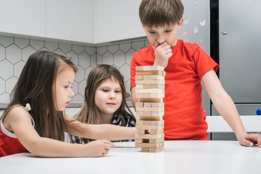Three children of concentrated thoughtful girls and boy playing wooden brick tower game on table. Balance challenge and moving blocks out of tower line. Construct skills and creativity games at home