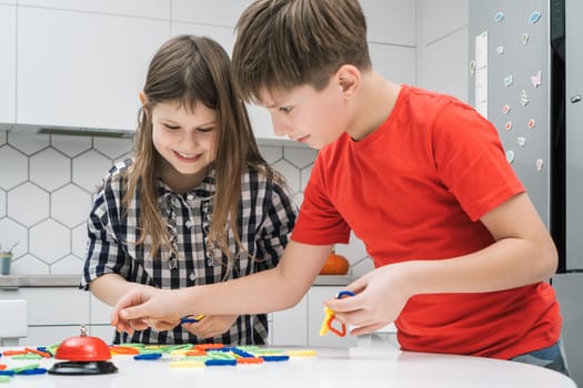 Laughing, funny boy and girl playing plastic chain game, connecting pieces together and making shape for time on white table in kitchen. Children creativity and motor, memory skills development