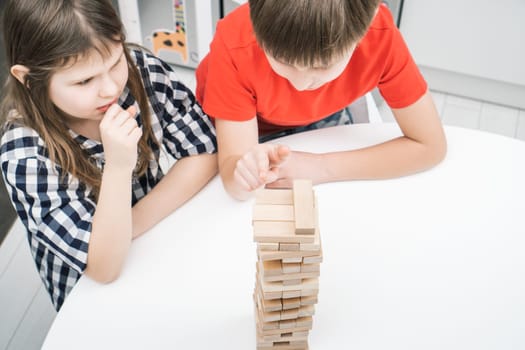 Serious, concentrated children, boy and girl thinking of strategy in board wooden brick tower game. Moving blocks carefully from pyramids on table. Smart and logical board games. High angle view