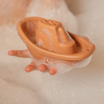 Unrecognizable child holding cute orange small silicone toy boat with holes on top with hand in bubble bath. Baby toys, toys for developing creativity, water games concept. Instagram use, square frame