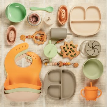 Pastel silicone set of tableware, cutlery, bibs, accessories and wooden toys for children on white cloth background. Baby accessories, tableware concept. Flat lay, top view, knolling, instagram use.