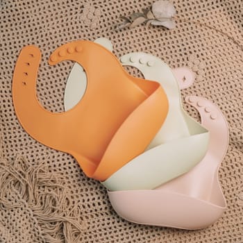 Three silicone baby bibs of different colors on beige blanket. Flat lay. Square frame instagram use