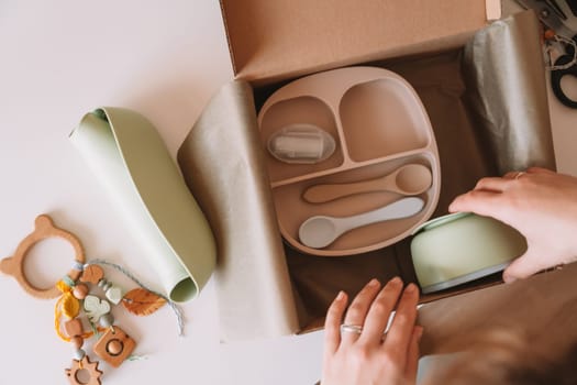 Cropped woman hands packing and wrapping shipment with silicone baby dish, plate, spoons, bowl, bib, wooden eco teether pacifier with trinket decoration, in cardboard box for delivery. Top view