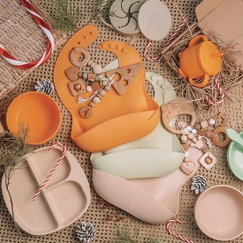 Set of silicone baby dishes, bibs and wooden teethers in beige shades on beige blanket with Christmas decorations. Flat lay. Square frame instagram use
