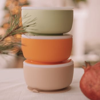 Set of silicone baby dishes. Three bowls with lids stacked on top of each other with pomegranate against background of Christmas tree. Creative blur and grain. Square frame instagram use