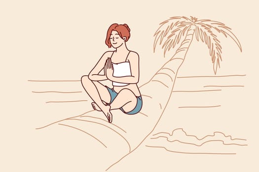 Woman doing yoga sitting on palm tree in lotus position enjoying summer travel and meditating on beach. Girl relaxing on beach with sea is fond of yoga and sunbathing during trip to island in ocean