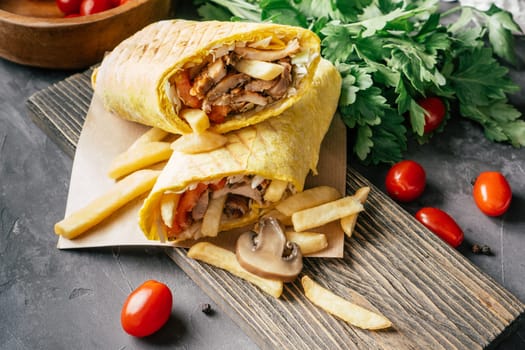 Rustic roll of shawarma with french fries, mushrooms, chicken and vegetables in pita bread on wooden dark background with cherry tomatoes and green parsley