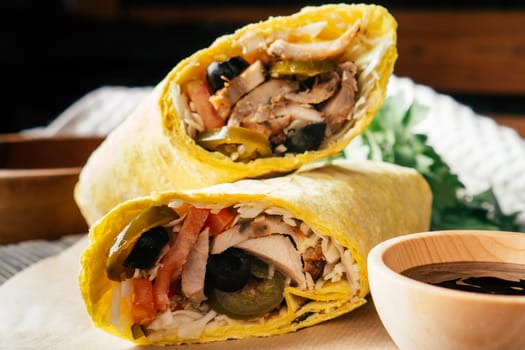 Mexican roll of shawarma with hot sauce, jalapeno pepper, olives, chicken and vegetables in pita bread on wooden dark background with cherry tomatoes and green parsley