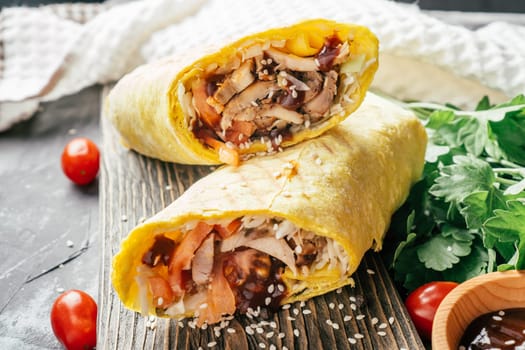 Roll of shawarma with sesame seeds, barbecue sauce, chicken and vegetables in pita bread on wooden dark background with cherry tomatoes and green parsley
