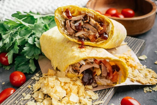 Crispy roll of shawarma with chips, barbeque sauce, chicken and vegetables in pita bread on wooden dark background with cherry tomatoes and green parsley