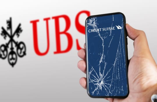 New York, US, March 2023: Hand holding a phone with Credit Suisse logo on cracked screen. UBS logo blurred on white background. UBS bought Credit Suisse for 3B CHF in stock. Illustrative editorial