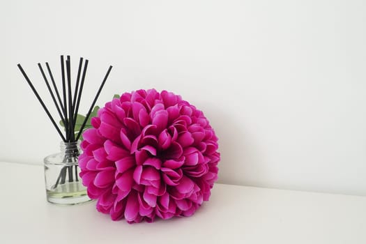 Arrangement with incense sticks, essential oil in a glass vase and a bright pink-lilac terry artificial flower in the interior of a white room. White background.