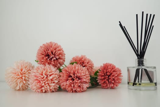 Composition with incense sticks, essential oil in a glass vase and pink terry artificial flowers in the interior of a white room. White background. Lots of flowers and scent sticks