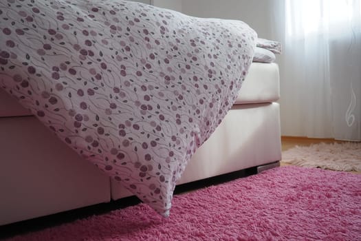 Cozy compact bedroom, white furniture, pink carpet and tulle curtains. Fluffy feather blanket thrown over the double bed. Romantic morning. Light from the window. The blanket hangs down to the floor