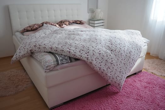 Cozy compact bedroom with white furniture pink carpet and tulle curtains. Fluffy feather blanket is thrown over the bed. Romantic morning. Double bed with upholstered headboard. Light from the window