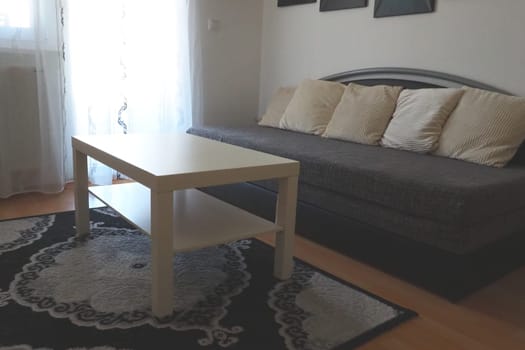 A white coffee table and a large gray sofa with decorative white cushions in the living room of a trendy apartment. Translucent tulle on the window or door from the balcony. Laminate and carpet
