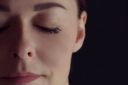 Close-up of half-face of pretty young woman with closed eyes with extreme long eyelashes, black eyeliner, ideal skin, perfect eyebrow on black background. Macro. Eyelash extension, beauty procedures.