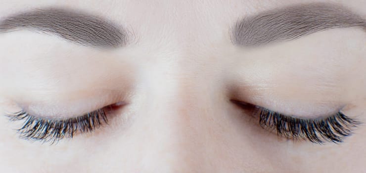 Close-up of closed eyes with long false eyelashes, black eyeliner, flawless skin, gorgeous eyebrows of unrecognizable young woman. Macro shot. Beauty procedures, cosmetology, microblading, make-up.