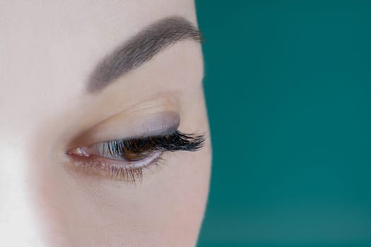 Close-up of brown eye with long eyelashes black eyeliner flawless eyebrow of unrecognizable young woman looking down on green background. Macro. Eyelash extension, cosmetology, make-up, advertisement.