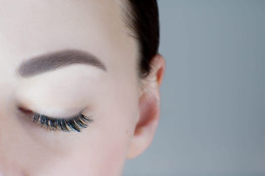 Close-up of closed eye with long eyelashes, black eyeliner, perfect skin, ideal eyebrow of unrecognizable young woman on grey background. Macro. Eyelash extension, beauty procedures, visage, make-up.