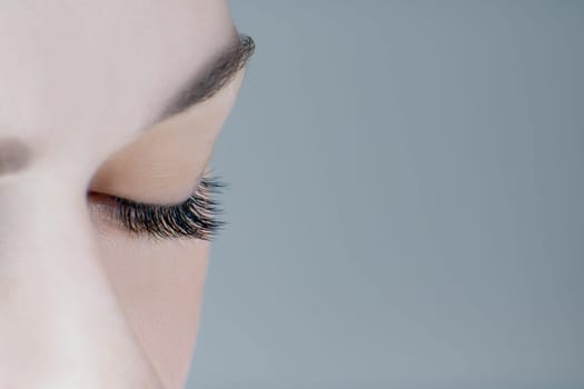 Close-up of closed eye with extreme long eyelashes, healthy skin, perfect eyebrow of unrecognizable young woman on grey background. Macro. Eyelash extension, beauty procedures, make-up, microblading.
