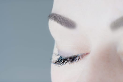 Close-up of closed eye with long artificial eyelashes, amazing skin, ideal eyebrow of unrecognizable young woman on grey background. Macro. Eyelash extension, beauty procedures, make-up, microblading.