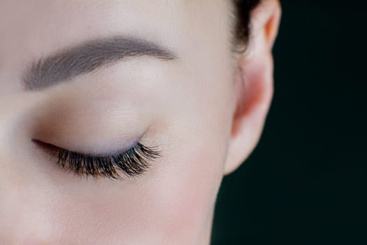 Close-up of closed eye with amazing long eyelashes, black eyeliner, perfect skin, eyebrow of unrecognizable young woman on black background. Macro. Eyelash extension, beauty procedures. Soft focus.