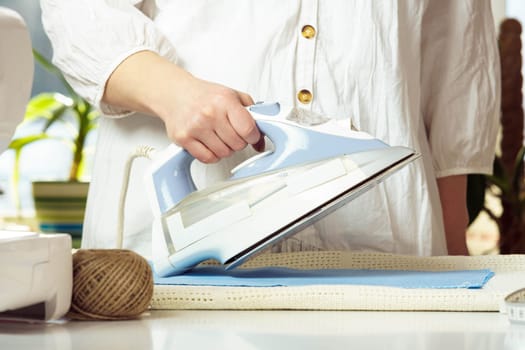 Cropped female in white shirt ironing, using iron steamer for blue wrinkled textile on table. Dressmaker prepare fabric, manufacturing work. Making clothes, unfolding textile. Small business and hobby