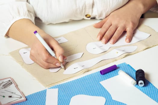 Cropped female seamstress drawing contours with pen of pinned paper sewing patterns on textile sheet on table. Needlework, making patterns for future items such as toys or clothes. Small business.
