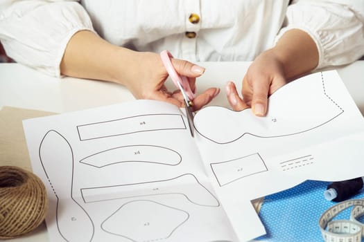 Close up unrecognizable female designer or tailor cutting patterns and shapes from paper for future textile stuffed toys in workshop. Handcraft, hobby diy master class. Needlework lessons in school