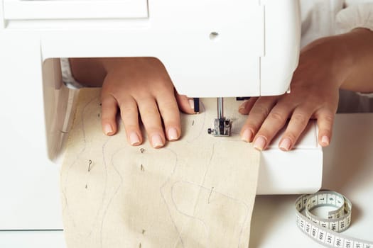 Close up hands of cropped woman dressmaker using sewing machine, place white fabric with stitch contour on needle plate under presser foot near tape measure in workshop. Production of clothes, toys