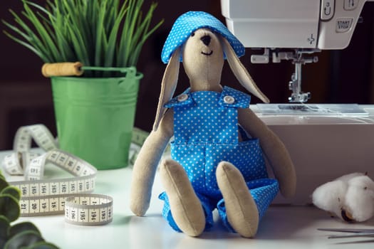 Close up cute, funny sewed rabbit toy in blue design costume sitting near houseplant, measure tape and cotton, leaning to sewing machine on table. Doll making for children, animal toys craft at home