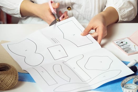 Cropped image of female designer employer hands cutting out patterns from paper sheet with numbers to make clothes and toys. Handcraft and needlework. Manufacture working in workshop, atelier