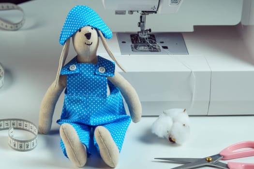 Close up funny smiling sewed stuffed rabbit toy in blue design costume, cocked hat, sit near measuring tape, cotton, scissors, next to sewing machine. Master class, tutorial, how to stitch animal doll
