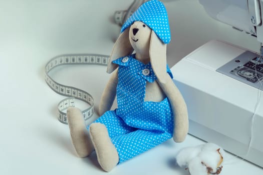 Close up cute, funny smiling sewed stuffed rabbit toy in blue design costume sitting near measure tape and cotton, leaning to sewing machine on table. How to stitch the toy, master class, tutorial