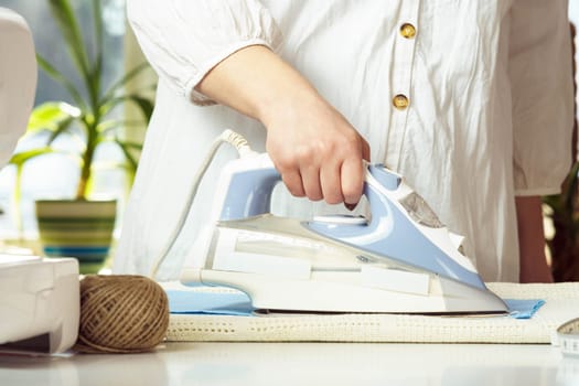 Cropped woman wearing white shirt steam iron blue wrinkled textile on table neat thread yarn. Dressmaker prepare fabric for sewing, manufacturing work indoors. Small business or hobby. Close up