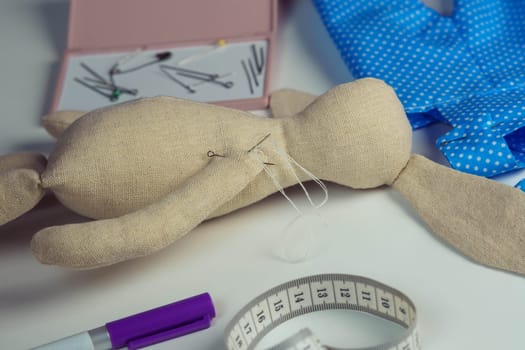 Sewing process of stuffed rabbit toy with needles in body lying on table. Knitting hobby. Hare toy near blue polka dot textile near measure tape and marker. Toys shop, store. DIY, children present