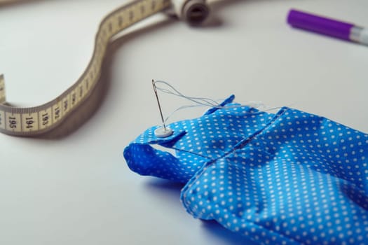 Small cute started blue polka dot costume with needle overalls for toy near measure tape and pen on white table. Sewing process of stuffed toys and clothes. Knitting hobby. DIY, children store