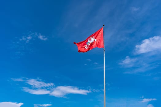 The historic flag of Lithuania, a rider with a sword on a white horse on a red background flutters against a bright blue sky with white clouds.