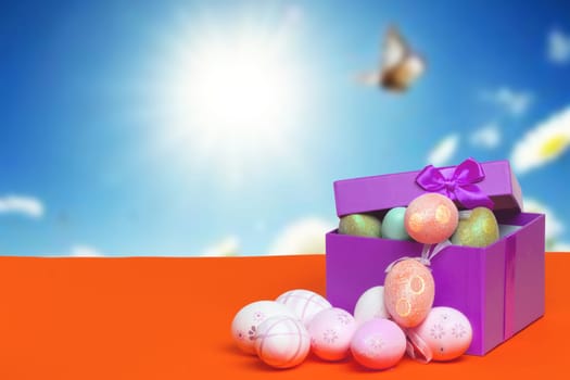 Happy Easter greeting card template. Close up of colourful Easter eggs and an open gift box with eggs on orange table against abstract blurred sunny sky background. Copy space for design.
