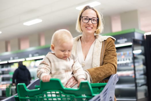 Caucasian mother shopping with her infant baby boy child choosing products in department of supermarket grocery store