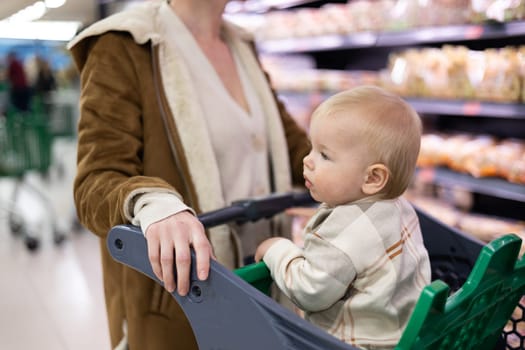 Caucasian mother shopping with her infant baby boy child choosing products in department of supermarket grocery store