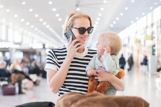 Mother talking on mobile phone while traveling with child, holding his infant baby boy at airport terminal waiting to board a plane. Travel with kids concept