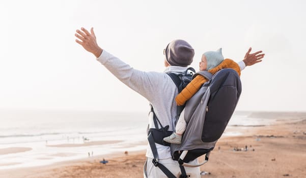 Young father rising hands to the sky while enjoying pure nature carrying his infant baby boy son in backpack on windy sandy beach. Family travel concept